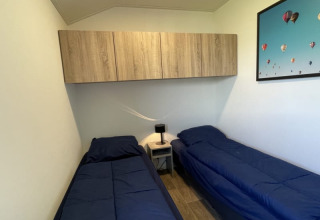 Kleines Zimmer in einem Tiny House mit zwei Einzelbetten, blauer Bettwäsche, Wandregal und Nachttisch.