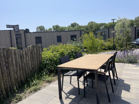 Buiten tafel en stoelen op het terras van een tiny house, omgeven door planten en onder blauwe hemel.