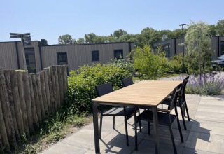 Mesa y sillas de exterior en la terraza de una tiny house, rodeada de vegetación y bajo un cielo azul.