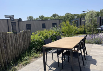 Udendørs bord og stole på terrasse ved tiny house, omgivet af grønne buske og træer under blå himmel.