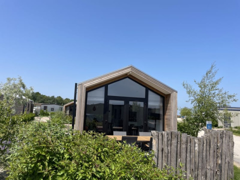 Een tiny house met grote ramen en houten omheining, omringd door groen onder een zonnige blauwe lucht.