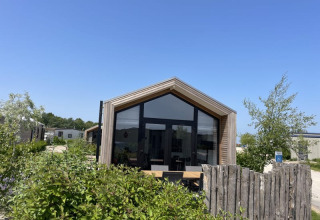 Een tiny house met grote ramen en houten omheining, omringd door groen onder een zonnige blauwe lucht.