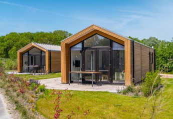 To moderne tiny houses med træfacader og store vinduer, omgivet af grønne planter og blå himmel.
