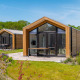 To moderne tiny houses med træfacader og store vinduer, omgivet af grønne planter og blå himmel.