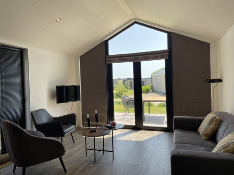 Lichte, moderne tiny house woonkamer met grote ramen, stoelen, zetel en zicht op een groene tuin.