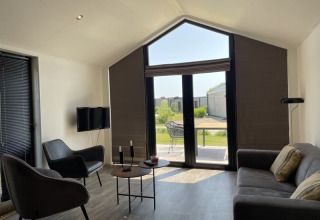 Lichte, moderne tiny house woonkamer met grote ramen, stoelen, zetel en zicht op een groene tuin.