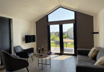 Lichte, moderne tiny house woonkamer met grote ramen, stoelen, zetel en zicht op een groene tuin.