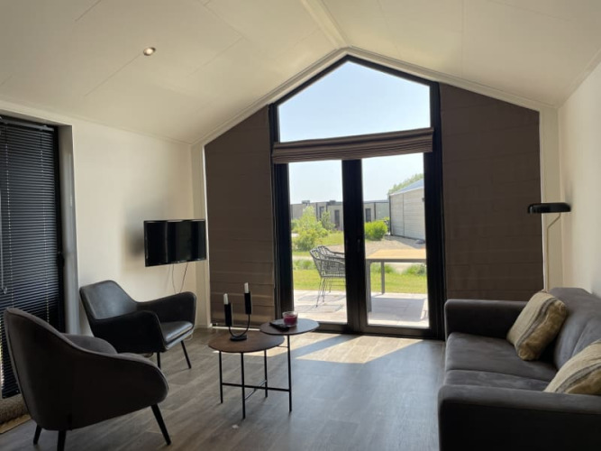 Luminoso salón moderno de tiny house con grandes ventanales, sillones, sofá y vistas a un jardín verde.