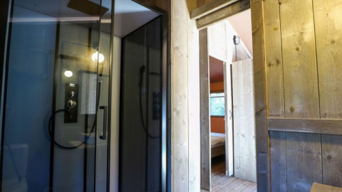 Vista interior de una cabaña con paneles de madera, cabina de ducha de vidrio y dormitorio al fondo.