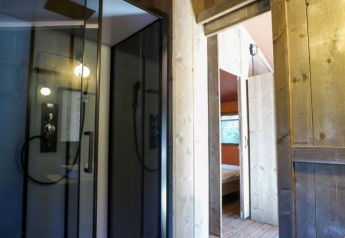 Vista interior de una cabaña con paneles de madera, cabina de ducha de vidrio y dormitorio al fondo.