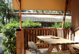 Terraza de madera con mesa de picnic en una cabaña, rodeada de arbustos verdes y árboles a la sombra.