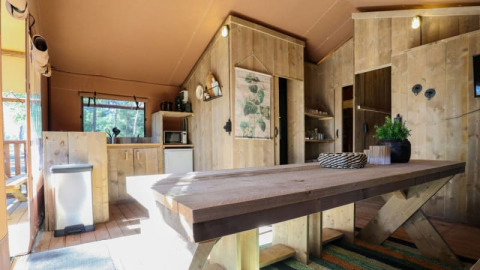 Interior de un lodge rústico con muebles de madera, mesa grande, cocina pequeña y luz natural.