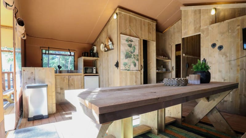 Interior de un lodge rústico con muebles de madera, mesa grande, cocina pequeña y luz natural.