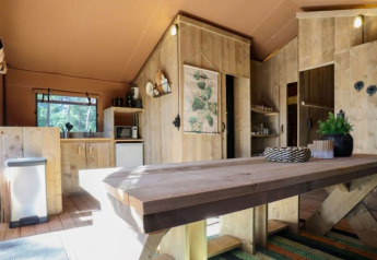 Interior de un lodge rústico con muebles de madera, mesa grande, cocina pequeña y luz natural.