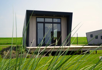 Modern tiny house with large windows and an angled roof, viewed through tall grass on a green lawn.