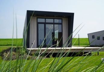Moderne tiny house med store vinduer og skråt tag på en grøn eng, set gennem højt græs i forgrunden.