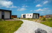 Een grindpad leidt naar moderne tiny houses omgeven door gras onder een heldere blauwe lucht met wolken.