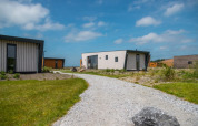 Un camino de grava conduce a modernas tiny houses rodeadas de césped verde bajo un cielo azul con nubes.