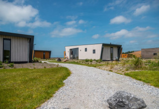 Een kiezelpad loopt naar moderne tiny houses, omringd door gras en een blauwe lucht met wolken.