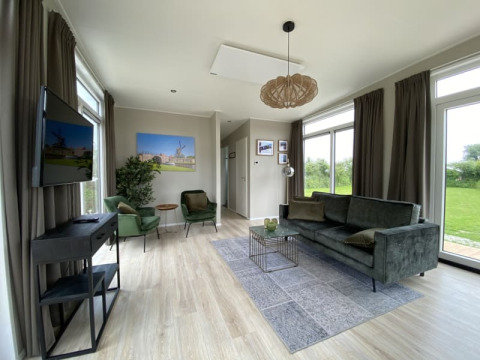 Sala luminosa y moderna de tiny house con sofá verde, sillones, ventanales y vista al jardín.