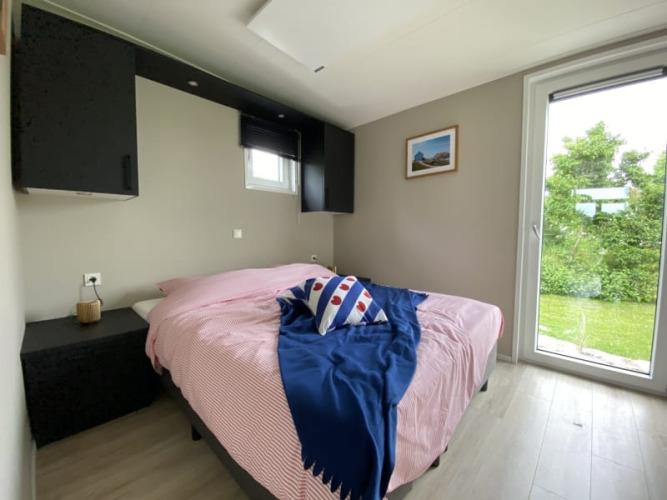 Dormitorio luminoso de tiny house con cama doble, gran ventana y vistas a un jardín verde exterior.