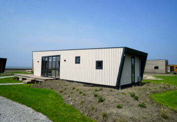 Modern tiny house with angular roof, large windows, and light wood siding on a grassy plot under blue sky.