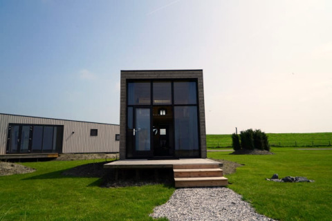 Lodge moderne avec de grandes fenêtres, terrasse en bois et pelouse verte sous un ciel bleu clair.