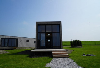 Lodge moderno con grandi finestre, veranda in legno e prato verde sotto un cielo azzurro limpido.