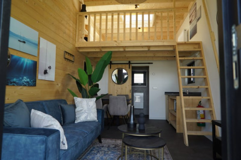Acogedora cabaña moderna con paredes de madera, altillo, sofá azul y pequeña cocina equipada.