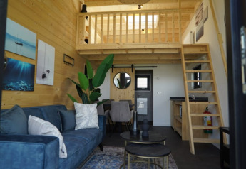 Acogedora cabaña moderna con paredes de madera, altillo, sofá azul y pequeña cocina equipada.