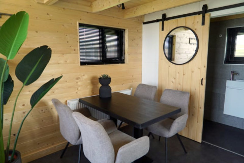 Salle à manger moderne de lodge avec bois clair, fenêtres noires, chaises beiges, table noire et grande plante.