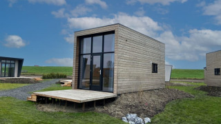 Moderni lodge in legno con grandi finestre, situati in un'area verde sotto un cielo azzurro e nuvoloso.