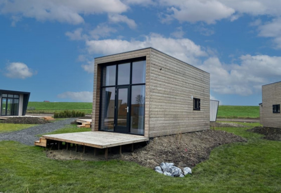 Moderni lodge in legno con grandi finestre, situati in un'area verde sotto un cielo azzurro e nuvoloso.