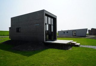 Lodge moderno in legno scuro con grandi finestre e veranda, situato su un prato verde sotto un cielo limpido.