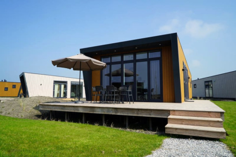 Lodge moderne avec terrasse en bois, mobilier d'extérieur et parasol, sur pelouse verte avec lodges voisins.