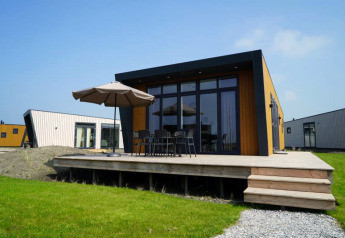 Modern lodge with wooden deck, outdoor furniture and umbrella, set on green grass with other lodges nearby.