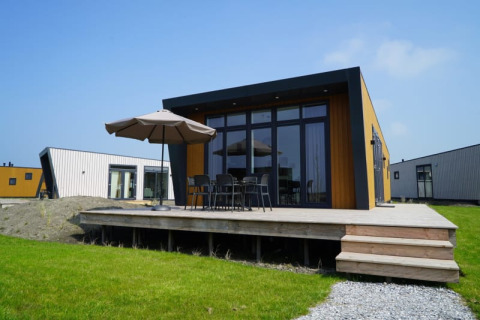 Lodge moderno con terraza de madera, muebles de exterior y sombrilla, rodeado de césped y otras cabañas.