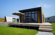 Lodge moderno con terraza de madera, muebles de exterior y sombrilla, rodeado de césped y otras cabañas.