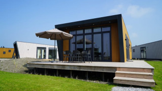 Lodge moderno con terraza de madera, muebles de exterior y sombrilla, rodeado de césped y otras cabañas.