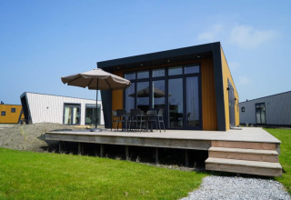 Lodge moderno con terraza de madera, muebles de exterior y sombrilla, rodeado de césped y otras cabañas.