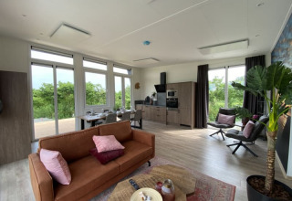 Intérieur de lodge moderne et lumineux avec cuisine ouverte, salon et grandes fenêtres sur la verdure.