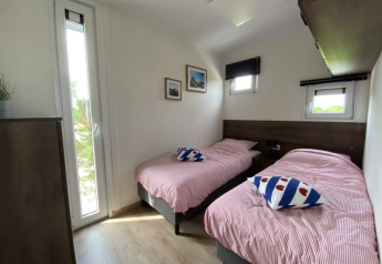 Chambre lumineuse dans un lodge avec deux lits simples, couettes rayées et fenêtres donnant sur la verdure.