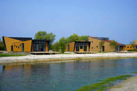 Moderne lodges met houten gevels aan een meer, omringd door bomen onder een heldere blauwe lucht.