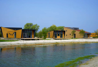 Modern lodge cabins with wood exteriors by a lakeside, surrounded by trees and blue daytime sky.