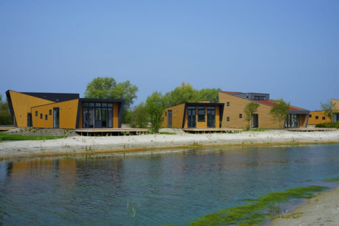 Cabañas modernas de madera junto a un lago, rodeadas de árboles y bajo un cielo azul durante el día.