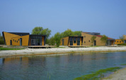 Cabañas modernas de madera junto a un lago, rodeadas de árboles y bajo un cielo azul durante el día.