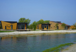Cabañas modernas de madera junto a un lago, rodeadas de árboles y bajo un cielo azul durante el día.
