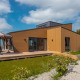 Moderne lodge med træbeklædning, stor terrasse og have under en blå himmel med spredte skyer.