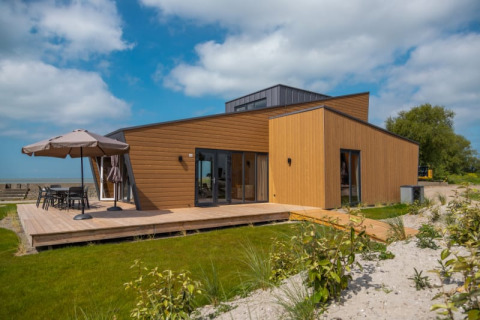 Lodge moderne en bois avec grande terrasse et jardin, sous un ciel bleu partiellement nuageux.