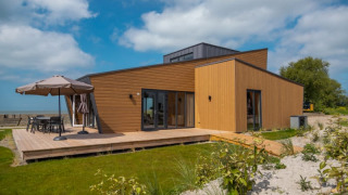 Moderne lodge med træbeklædning, stor terrasse og have under en blå himmel med spredte skyer.